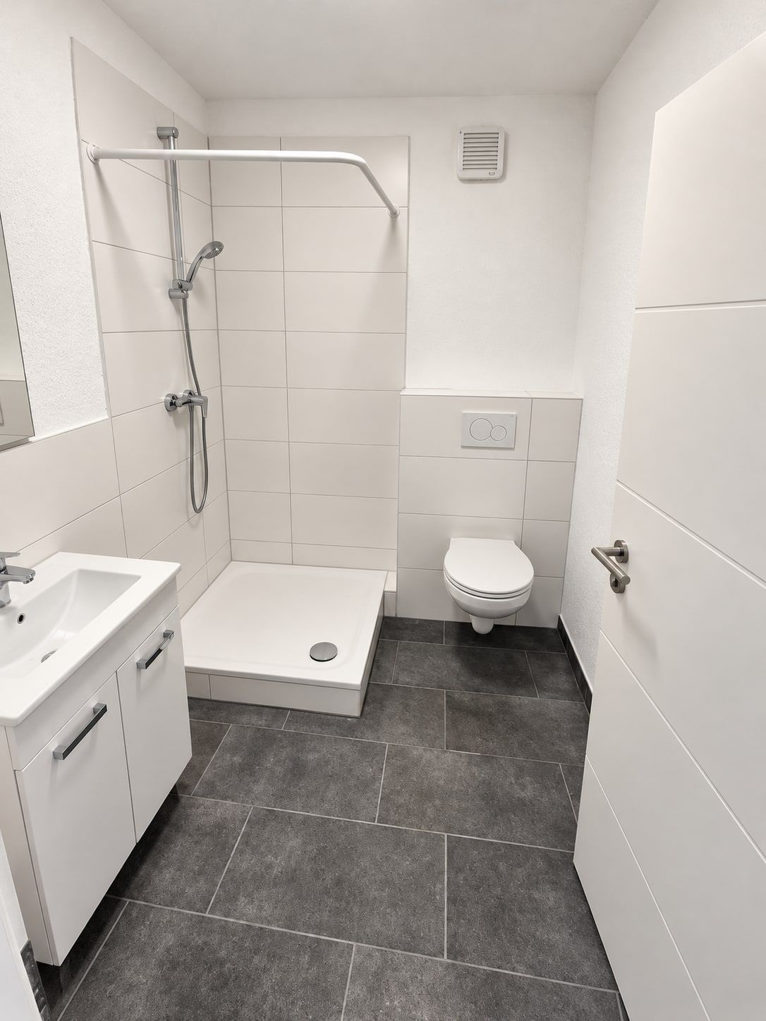 Bad von der Tür aus: Waschtisch mit Unterschrank, Dusche mit Duschwanne und Stange für den Vorhang, Wand-WC, weiße Wandfliesen und dunkler Fliesenboden.