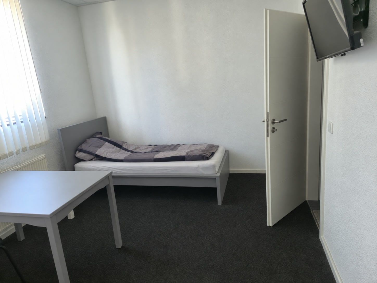 Einzelbett an der Wand, vorne der Tisch, rechts der Fernseher an der Wand neben der offenen Tür zum Bad.