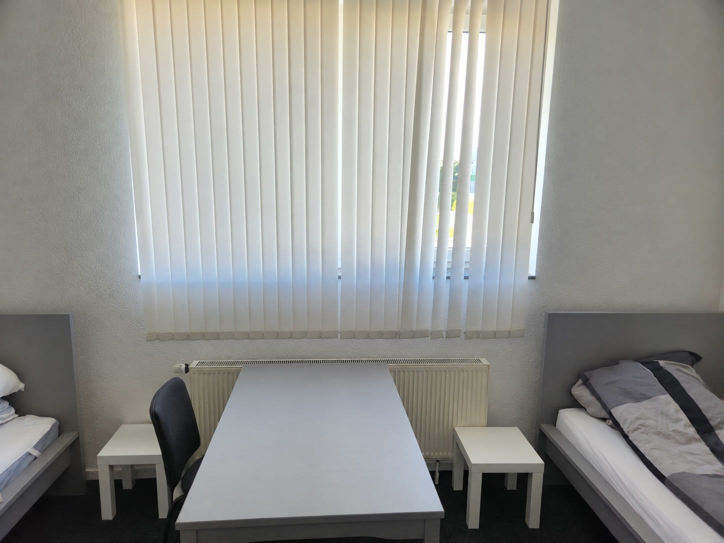 Grauer Tisch mit Stuhl vor dem Fensterband mit Lamellenvorhängen, links und rechts je ein Einzelbett, dunkler Teppich.
