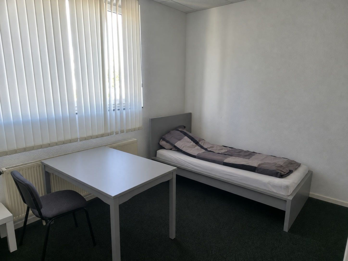 Tisch mit Stuhl und Einzelbett unter dem Fenster mit Lamellenvorhang und Heizkörper.