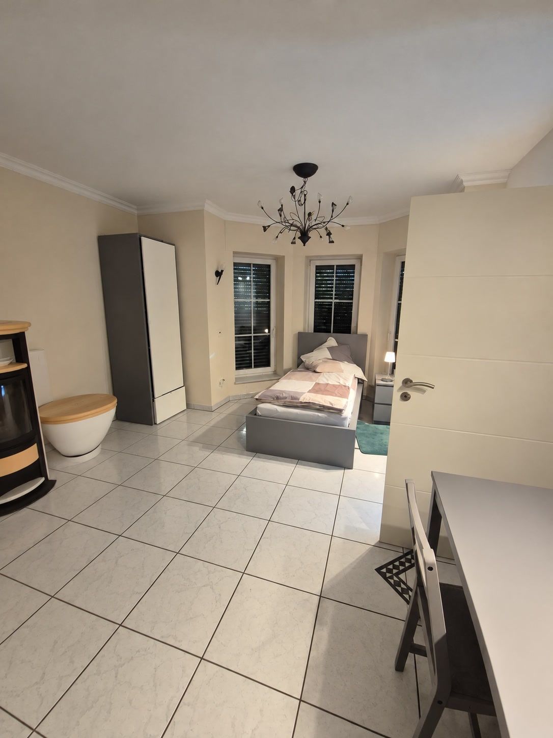 Blick vom Eingang: helle Fliesen, Kronleuchter, Kleiderschrank und Bett vor dem Erker mit Sprossenfenstern, links der Kaminofen.