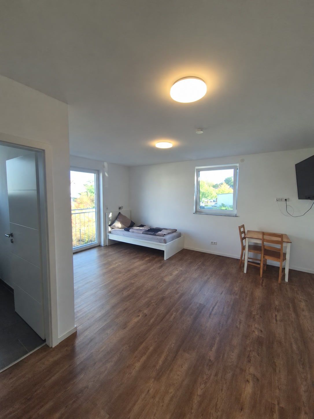 Gesamtansicht: Einzelbett vor der Fenstertür, Holztisch mit zwei Stühlen unter dem Fenster, Fernseher am Wandarm.