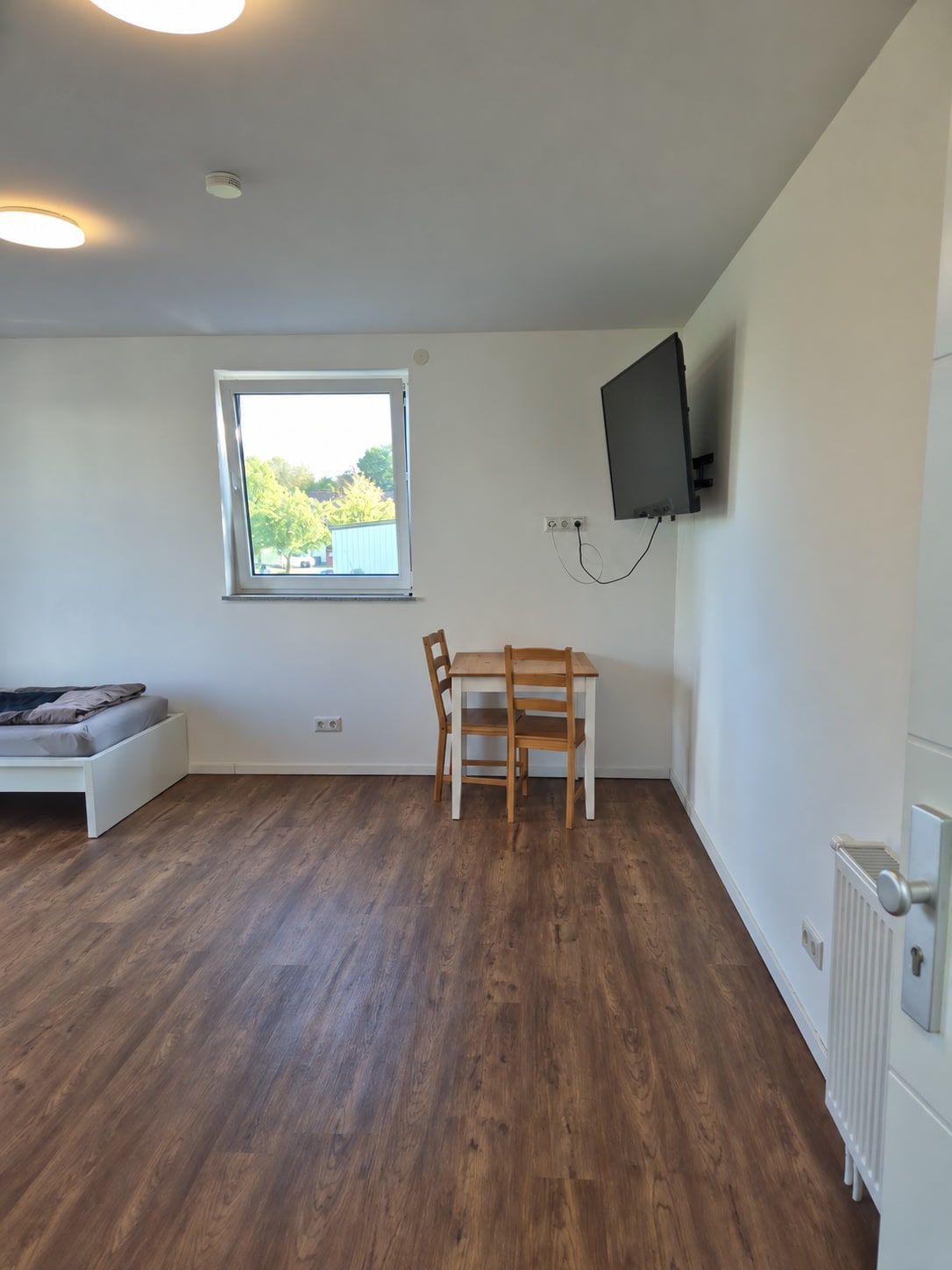 Holztisch mit zwei Stühlen unter dem Fenster, Fernseher am Wandarm, Heizkörper an der Wand.