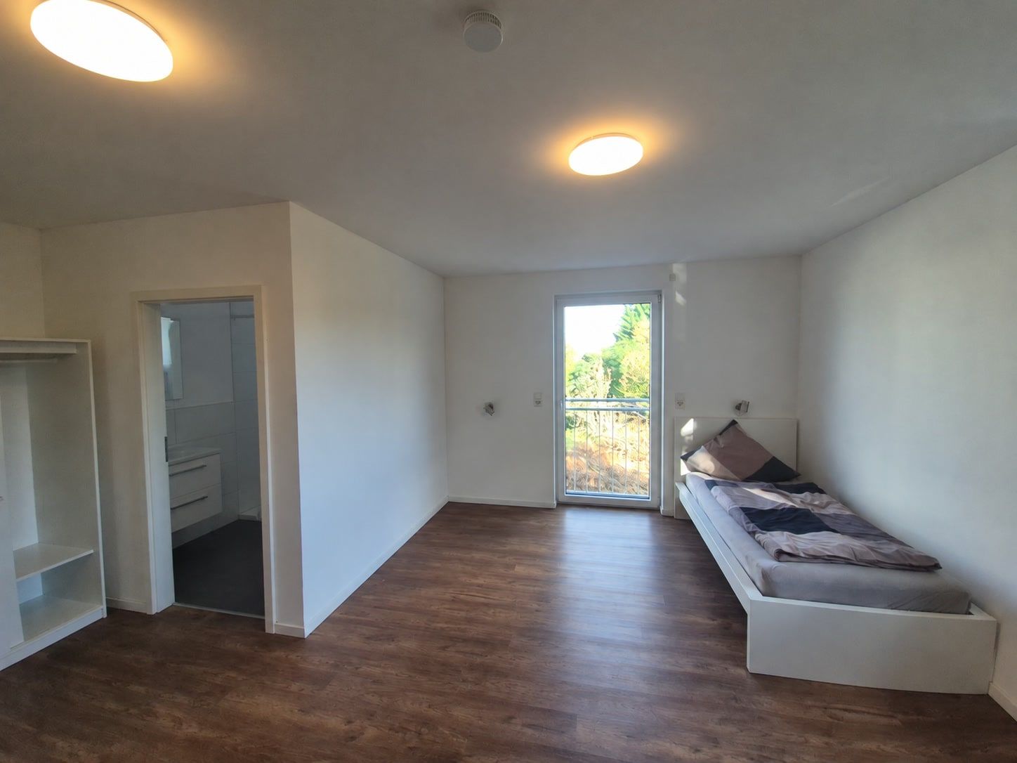 Heller Raum mit Holzboden: offener Schrank, Tür zum eigenen Bad, Einzelbett an der Wand und bodentiefe Fenstertür mit Geländer.